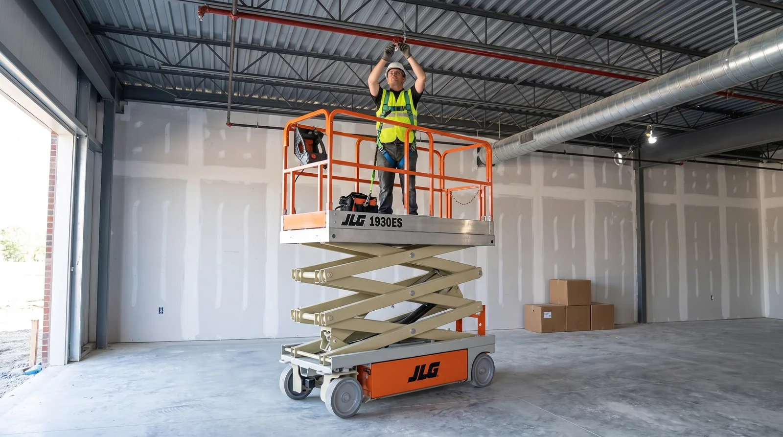An orange-railed electric scissor lift raised in an empty steel-framed commercial unit, a harnessed worker in a hard hat reaching up to overhead pipework beneath the roof deck.