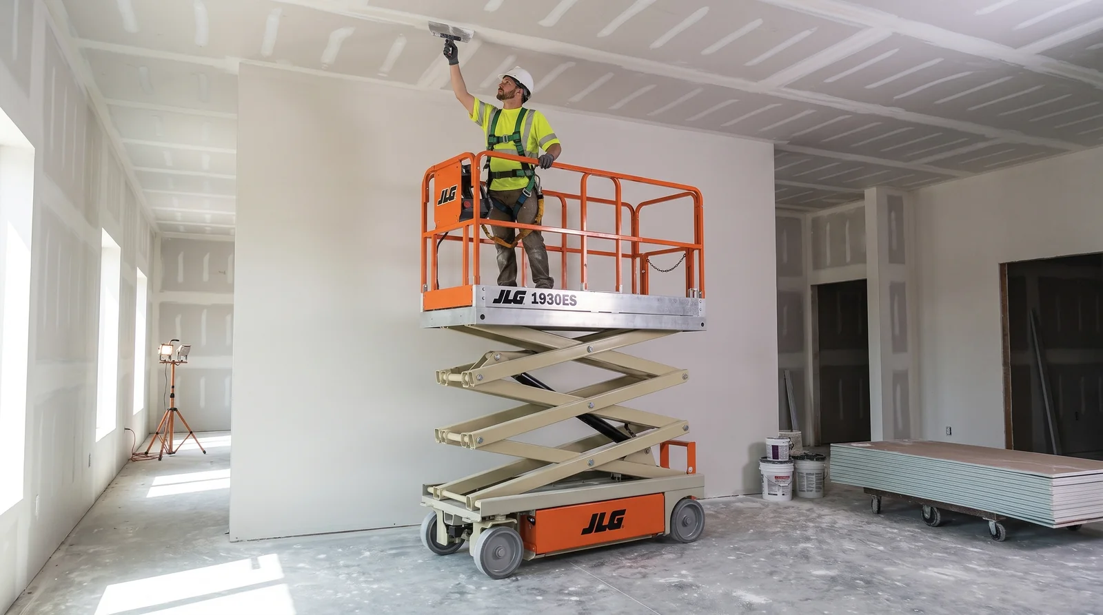 An electric scissor lift raised in a newly drywalled room, a harnessed worker reaching up to finish the taped ceiling seams, with a cart of drywall sheets and mud buckets on the floor.