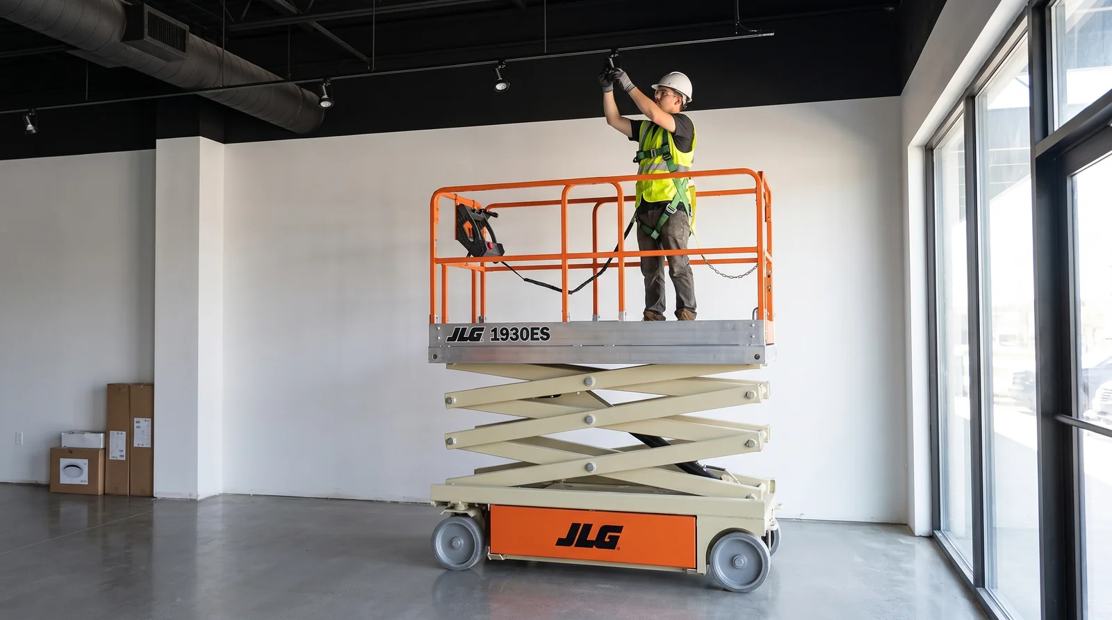 An electric scissor lift raised beside the storefront windows of a white-walled retail unit, a harnessed worker adjusting a track light on the black-painted ceiling.