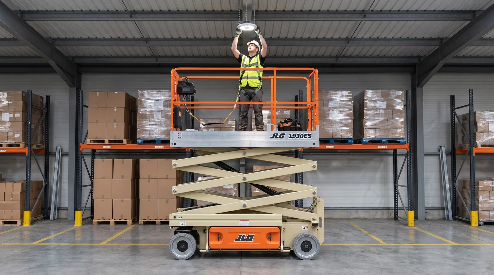 An electric scissor lift raised in a warehouse aisle in front of racking loaded with cartons, a harnessed worker reaching up to a hanging LED light fixture.
